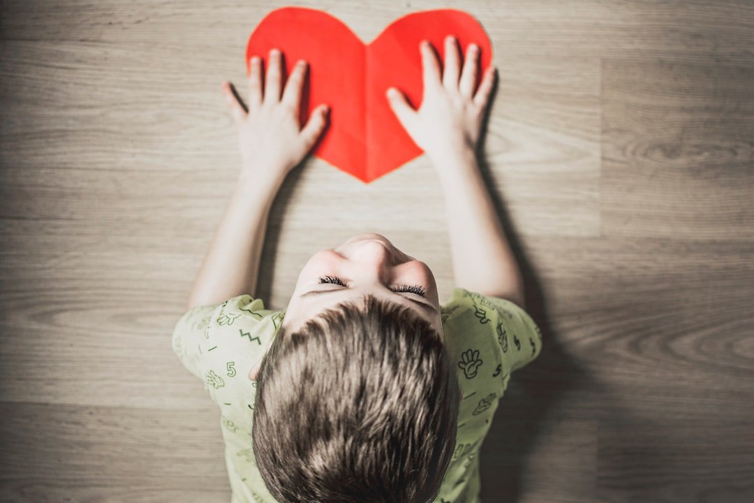Photo by Anna Kolosyuk boy in green shirt holding red paper heart cutout on brown table